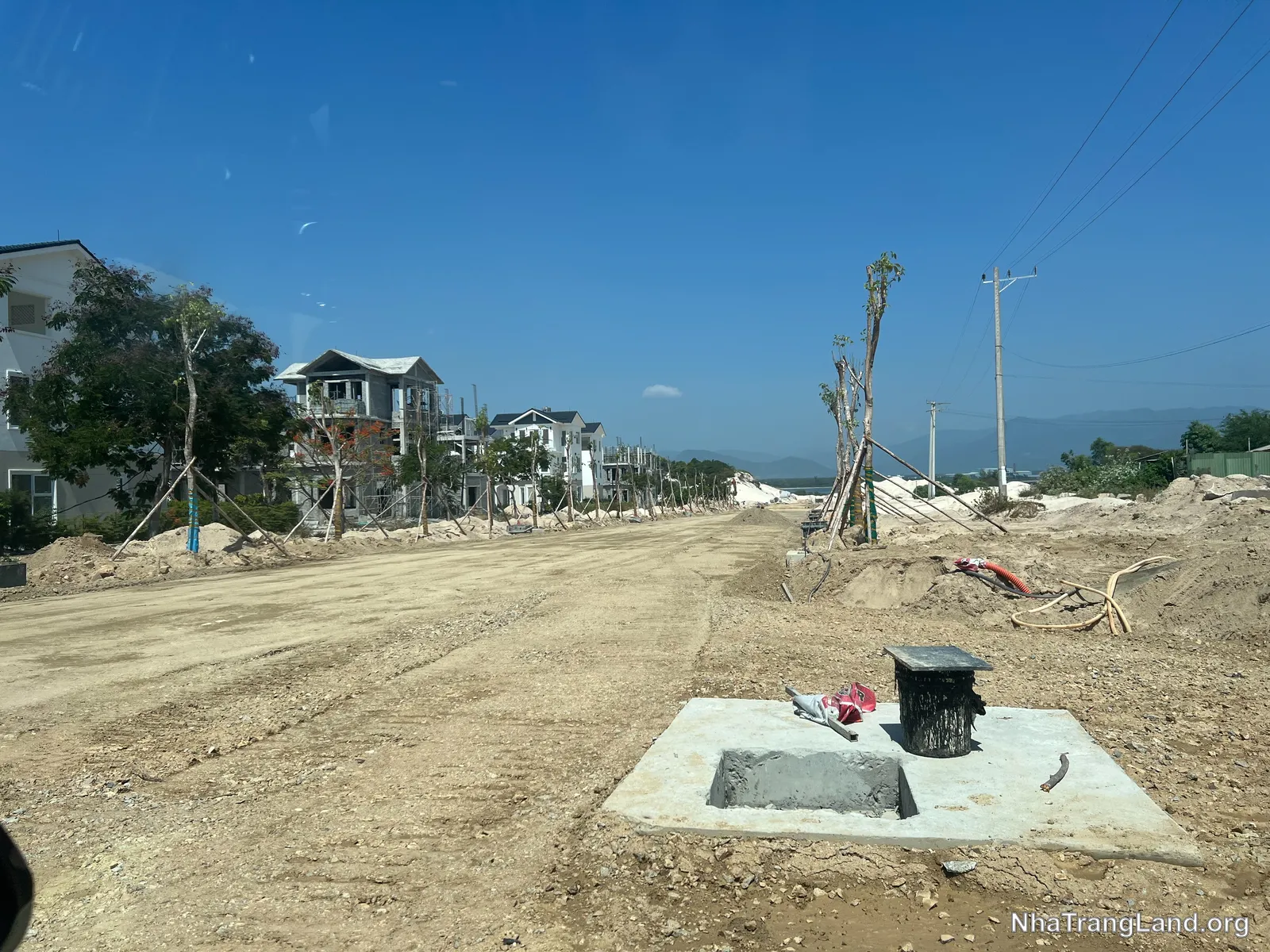 Tuyến đường nội khu mới: nền đường đã lu lèn, hố ga thoát nước hoàn thành, cây xanh trồng chống dựng hai bên — phía xa là dãy biệt thự đang xây thô.